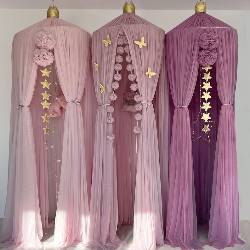 May include: Three pink and purple tulle canopy beds with gold stars and pom poms hanging from the top. The canopies are set up in a row against a white wall.