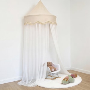 May include: A cream-colored canopy with a scalloped trim and fringe hangs from the ceiling. Sheer white curtains drape down, framing a white rocking chair with a stuffed bunny and a white rug with stuffed animal toys.