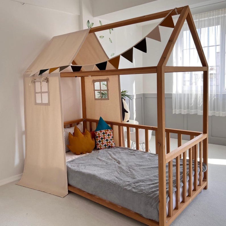 May include: A wooden house-shaped bed frame with a light-colored canopy and decorative pennant flags. The bed has a gray blanket, pillows, and a small window detail. The frame is made of natural wood.