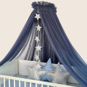 Può includere: Un baldacchino blu navy con la parte superiore a balze è sospeso sopra una culla. Ornamenti a forma di stella bianca pendono dal baldacchino. Cuscini a forma di stella argentati e blu e un lenzuolo con angoli blu completano l'arredamento della cameretta.