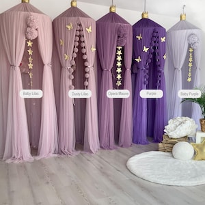 May include: Five different colored tulle canopy beds with gold star and butterfly decorations. The canopies are in shades of purple, lilac, and white. The canopies are hanging from the ceiling and are arranged in a row. The canopies are all the same size and shape. The canopies are all made of tulle and have a soft, flowing texture.