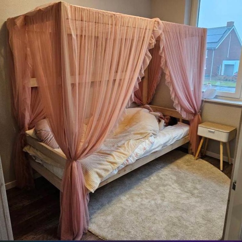 May include: A wooden bed frame with a pink canopy draped over it. The bed is made with white bedding and a pink pillow. The bed is in a bedroom with a white rug and a small wooden nightstand.