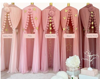 May include: Five hanging fabric canopies in shades of baby pink, pink, dusty pink, blush, and dusty rose. Each canopy features a crown top, sheer draped fabric, pom-pom garlands, and gold butterfly or star accents with labels.