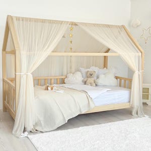 Peut inclure: Un cadre de lit en bois clair en forme de maison avec une structure de toit en pente drapée de rideaux en tissu crème transparent. Le lit est habillé de linge blanc, d'oreillers en forme de nuage et d'un ours en peluche, avec des étoiles dorées.