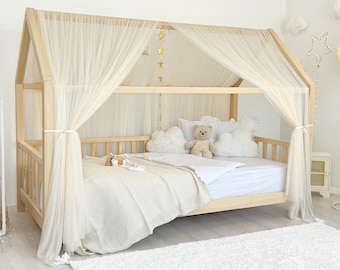 May include: A light-colored wooden house-shaped bed frame featuring a peaked roof structure draped with sheer cream-colored fabric curtains. The bed is dressed with white bedding, cloud-shaped pillows, and a teddy bear, accented by hanging gold star decor.
