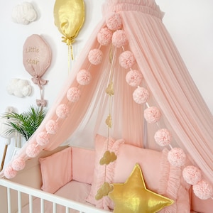 Puede incluir: Un dosel de tul rosa con una cuna blanca debajo. El dosel está decorado con una cadena de pompones rosas. La cuna tiene un parachoques rosa y una almohada rosa con una estrella dorada. También hay dos globos dorados y una nube blanca colgando de la pared.