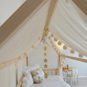 May include: A wooden house-shaped bed frame draped with sheer white fabric curtains and a garland of cream-colored pom-poms. A hanging mobile with gold stars is suspended from the center. A plush teddy bear and white pillows sit on the bed.
