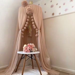 Könnte beinhalten: Ein Baldachin aus rosa Tüll mit weißem Pomponbesatz und einer Schnur aus rosa Pompons, die von oben hängen. Vor dem Baldachin steht ein kleiner weißer Tisch mit Holzbeinen, auf dem ein Strauß rosa Rosen liegt.