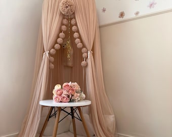 May include: A blush pink tulle canopy with a white pom-pom trim and a string of blush pink pom-poms hanging from the top. A small white table with wooden legs sits in front of the canopy, with a bouquet of pink roses on top.