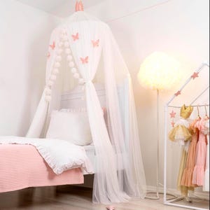 Può includere: Un letto a baldacchino bianco con una baldacchino in tulle bianco e decorazioni a farfalla rosa. Il letto ha un piumino bianco e una coperta rosa. C'è una lampada bianca con un paralume bianco sullo sfondo.
