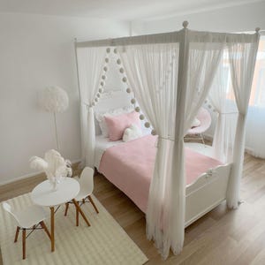May include: A white canopy bed with a pink blanket and white pom pom trim. The bed is in a room with white walls and a white rug.