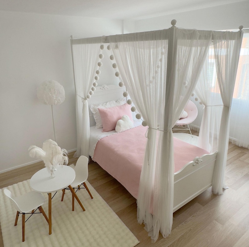 May include: A white canopy bed with a pink blanket and white pom pom trim. The bed is in a room with white walls and a white rug.