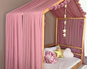 May include: A wooden house-shaped bed frame draped with dusty rose fabric curtains. The frame features a row of matching pom-poms along the roofline and a hanging garland of gold stars. Inside, a gold star-shaped pillow and a large pink flower.