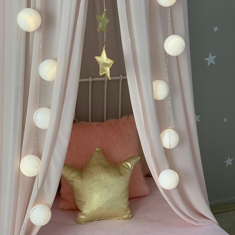 Puede incluir: Una cama con dosel rosa con una almohada dorada en forma de estrella y decoraciones colgantes de estrellas. Cadenas de luces blancas brillantes en forma de globo est&aacute;n drapeadas alrededor del dosel. Una almohada rosa esponjosa a&ntilde;ade textura.