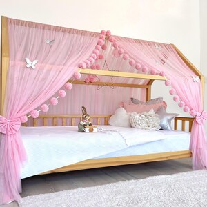 May include: A wooden framed bed with a pink canopy and decorative accents. The canopy is draped with sheer pink fabric and adorned with pink pom-poms, star-shaped lights, and butterfly decorations. The bed is made up with white bedding and pillows.