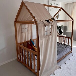 May include: A wooden house-shaped bed frame with a beige fabric roof and curtains. The bed has a low profile with a mattress and pillows. The frame is made of light-colored wood, and the design includes decorative elements.