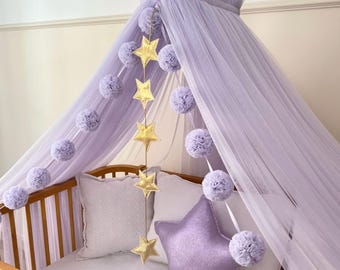 May include: A light purple tulle bed canopy featuring a hanging string of five metallic gold stars. The canopy is draped over a wooden crib containing a white pillow and a large, star-shaped purple pillow with a subtle glittery finish.