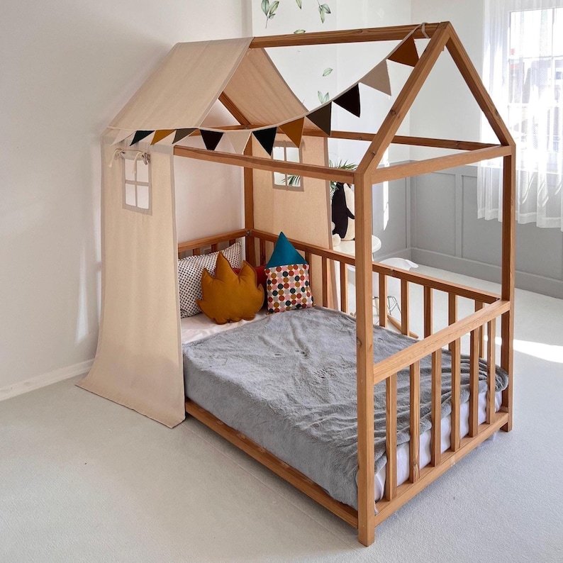 May include: A wooden house-shaped bed frame with a light-colored fabric roof and side panel. The bed is decorated with colorful pillows and a gray blanket. A garland of small flags hangs across the top. The bed is designed for children.