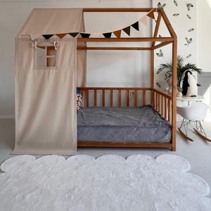 May include: A wooden house-shaped bed frame with a beige canopy and curtains. The bed has a gray blanket and a white cloud-shaped rug in front. A garland of brown, black, and beige triangles hangs above the bed.