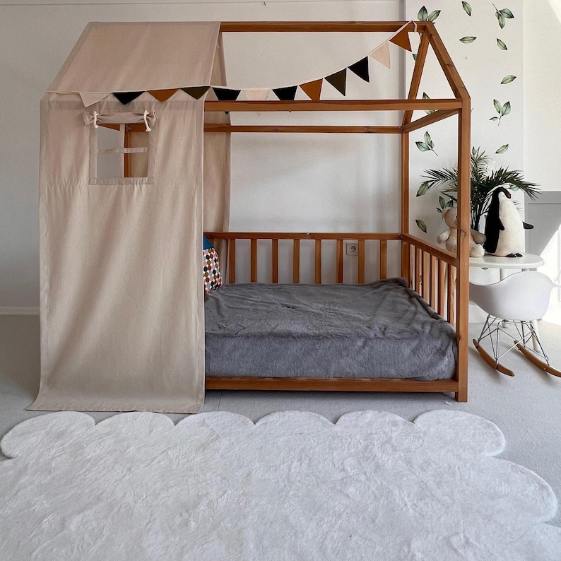 May include: A wooden house-shaped bed frame with a beige canopy and curtains. The bed has a gray blanket and a white cloud-shaped rug in front. A garland of brown, black, and beige triangles hangs above the bed.