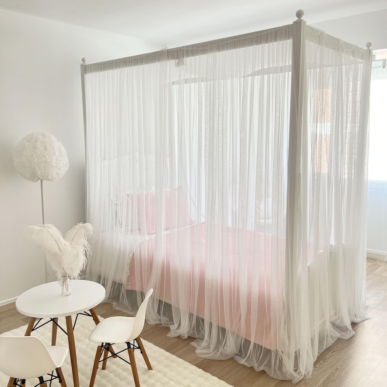 May include: A white canopy bed with a pink bedspread and white sheer curtains. The bed is in a bedroom with a white rug and two white chairs with wooden legs.
