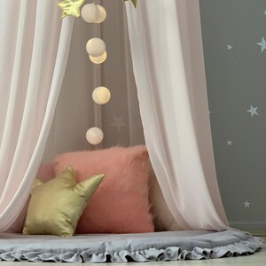 Puede incluir: Un dosel rosa con cortinas transparentes, una almohada dorada en forma de estrella y una almohada rosa esponjosa. Una cadena de globos blancos brillantes cuelga de la parte superior, y una alfombra gris con borde con volantes se encuentra debajo. El fondo es una pared gris con estrellas blancas.