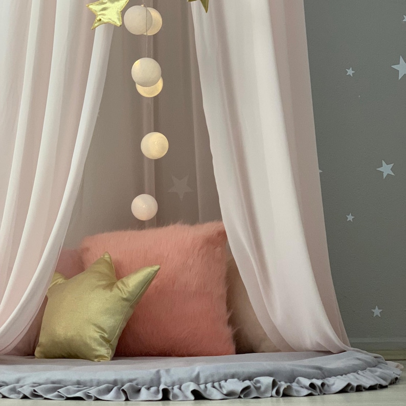 Puede incluir: Un dosel rosa con cortinas transparentes, una almohada dorada en forma de estrella y una almohada rosa esponjosa. Una cadena de globos blancos brillantes cuelga de la parte superior, y una alfombra gris con borde con volantes se encuentra debajo. El fondo es una pared gris con estrellas blancas.