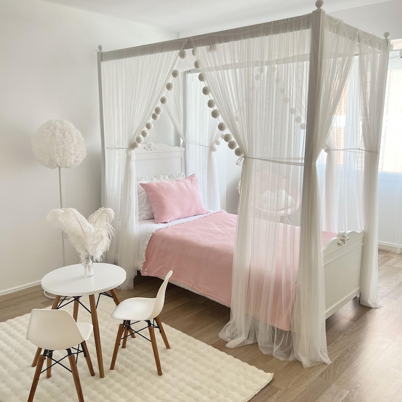 May include: A white canopy bed with a pink striped comforter and white sheer curtains. The bed is decorated with white pom poms and has a white wooden frame.