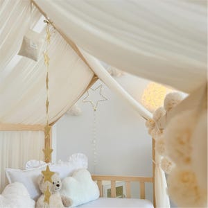 May include: A wooden house-shaped bed frame draped with sheer white fabric, featuring a hanging gold star garland, a light-up star ornament with a string of beads, and white cloud-shaped pillows surrounding a plush teddy bear.