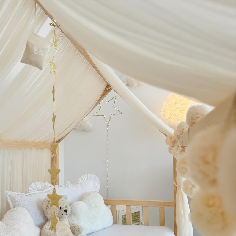 May include: A wooden house-shaped bed frame draped with sheer white fabric, featuring a hanging gold star garland, a light-up star ornament with a string of beads, and white cloud-shaped pillows surrounding a plush teddy bear.