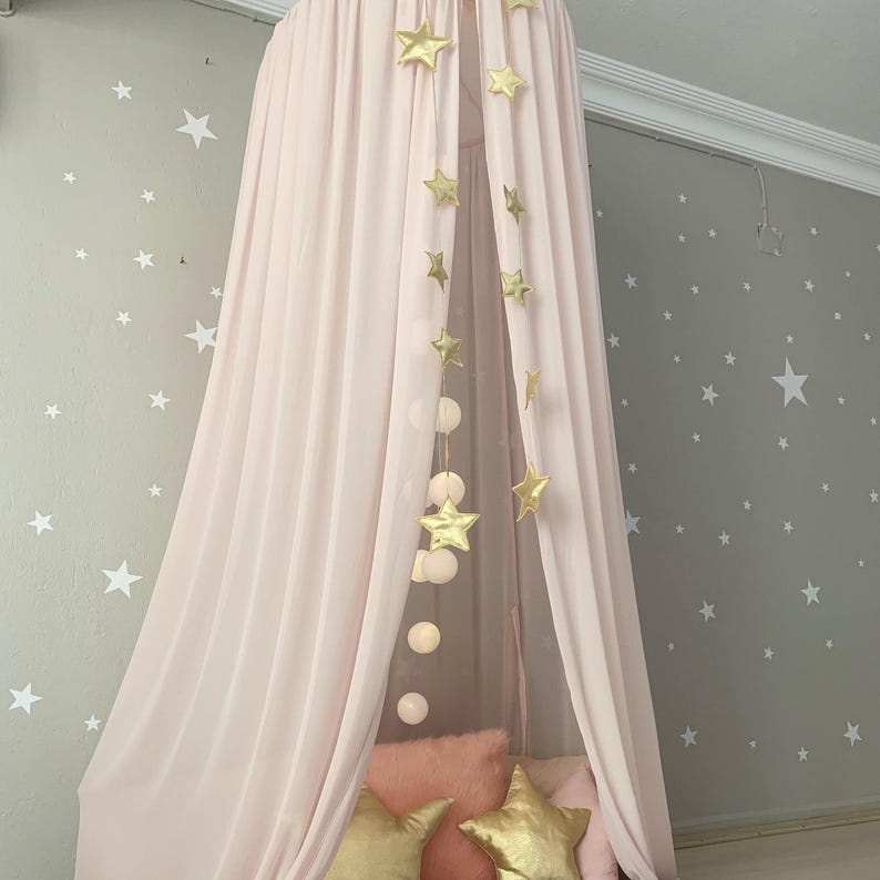Puede incluir: Una cama con dosel rosa con decoraciones de estrellas doradas y luces de cadena. La cama est&aacute; cubierta con tela transparente y tiene cojines rosas y dorados. La pared detr&aacute;s de la cama es gris con calcoman&iacute;as de estrellas blancas.