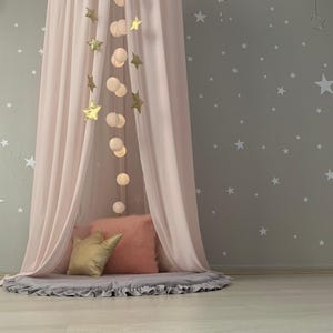 Puede incluir: Un dosel rosa con una alfombra gris con volantes y cojines decorativos. Luces doradas en forma de estrella y redondas cuelgan del dosel. La pared es gris con calcoman&iacute;as de estrellas blancas, creando un ambiente de guarder&iacute;a o dormitorio.