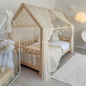 May include: A light-colored wooden house-shaped bed frame featuring a white tulle canopy draped over the roof structure. The bed includes a white mattress, a beige textured blanket, and a string of small pom-pom decorations hanging from the frame.