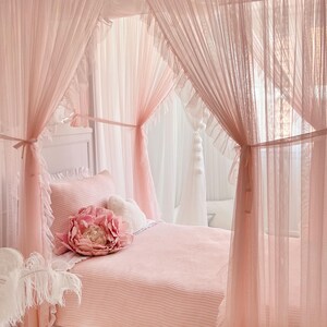 May include: A pink canopy bed with a white headboard and a pink and white striped comforter. A large pink flower pillow is on the bed. The canopy is made of sheer pink fabric with ruffles.