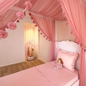 May include: A pink canopy bed with sheer curtains and a matching bedspread. The bed is decorated with pink pom-poms and a doll. The room has a white headboard and a small decorative house.