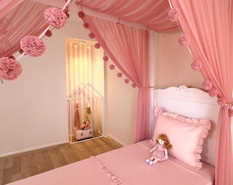 May include: A canopy bed featuring soft pink fabric drapes adorned with strings of matching pink pom-poms. The bed includes a pink ruffled pillow and a small doll sitting on the pink bedding. A white house-shaped clothing rack stands nearby.