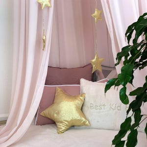 Puede incluir: Una acogedora cama con dosel rosa claro con decoraciones de estrellas doradas. La cama est&aacute; adornada con una almohada dorada en forma de estrella y otras almohadas en tonos rosa y blanco. La almohada blanca tiene bordado "Best Kid".