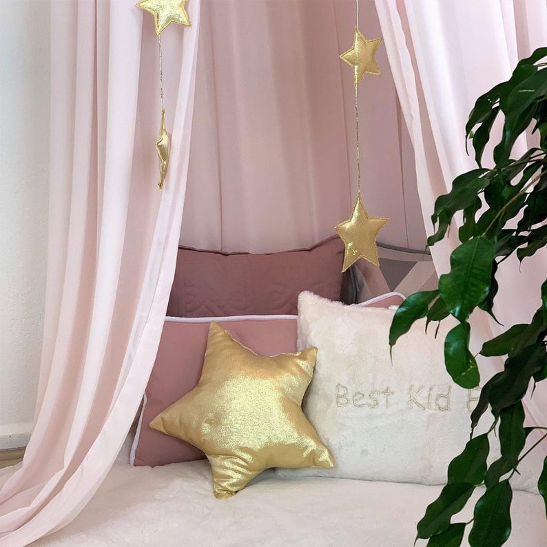 Puede incluir: Una acogedora cama con dosel rosa claro con decoraciones de estrellas doradas. La cama est&aacute; adornada con una almohada dorada en forma de estrella y otras almohadas en tonos rosa y blanco. La almohada blanca tiene bordado "Best Kid".