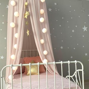 Puede incluir: Una cama de metal blanco con dosel y ropa de cama rosas. El dosel est&aacute; cubierto con tela rosa transparente y adornado con luces de cadena y decoraciones de estrellas doradas. La pared gris est&aacute; decorada con calcoman&iacute;as de estrellas blancas.