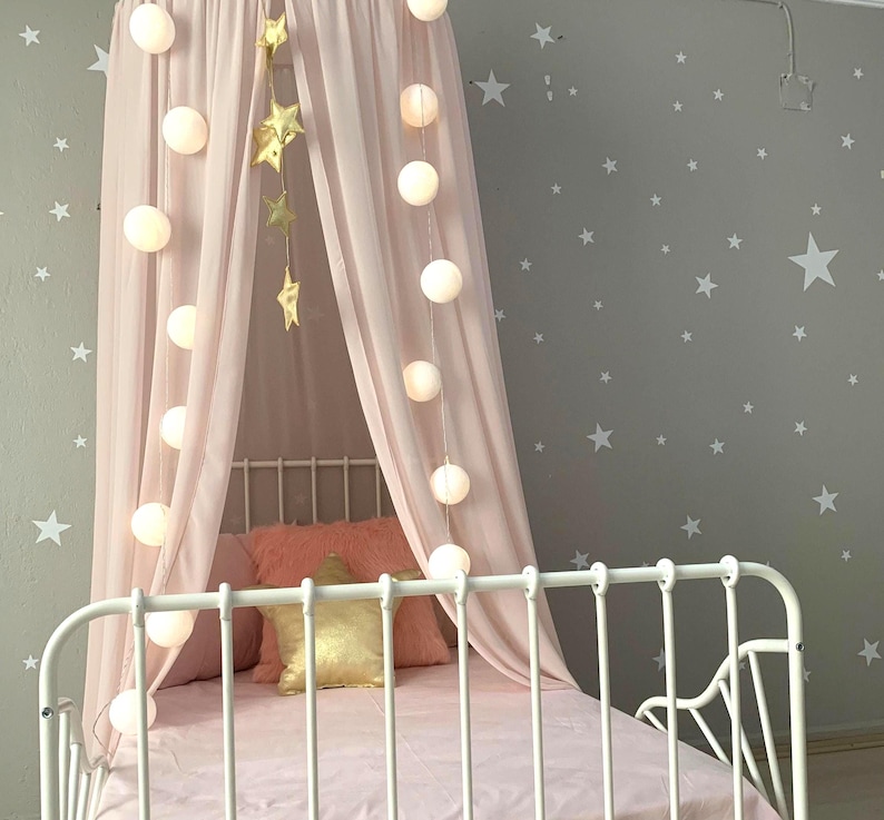 Puede incluir: Una cama de metal blanco con dosel y ropa de cama rosas. El dosel est&aacute; cubierto con tela rosa transparente y adornado con luces de cadena y decoraciones de estrellas doradas. La pared gris est&aacute; decorada con calcoman&iacute;as de estrellas blancas.