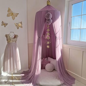 May include: A mauve tulle canopy with a white cushion and a gold star mobile hanging from the top. The canopy is set up in a room with a white wall and a window.