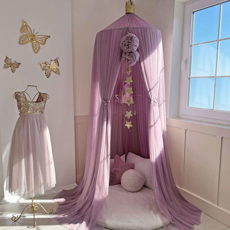 May include: A mauve tulle canopy with a white cushion and a gold star mobile hanging from the top. The canopy is set up in a room with a white wall and a window.