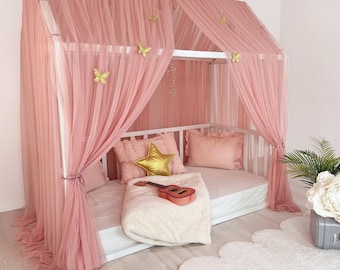 Op de afbeelding: Een wit huisvormig bedframe gedrapeerd met transparante oudroze tule gordijnen. De hemel is versierd met decoratieve gouden vlinders. Binnenin liggen roze kussens, een goudkleurig stervormig kussen, een crèmekleurige deken en een kleine houten speelgoedgitaar.