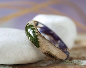 May include: A gold ring with a green leaf embedded in clear resin. The ring is resting on a white stone.