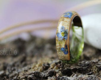 May include: A wooden ring with blue flowers embedded in the wood. The ring is on a mossy surface.