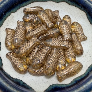 May include: A collection of small, amber-colored glass fish beads with detailed scale patterns. The beads are scattered inside a ceramic bowl with a blue and white rim, creating a textured and reflective display.