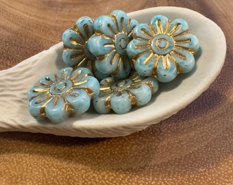 Puede incluir: Un grupo de seis cuentas de vidrio de color azul turquesa con detalles dorados en forma de flor. Las cuentas están dispuestas en una cuchara de cerámica blanca.
