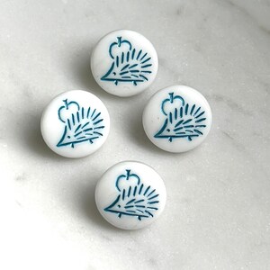 May include: Four round white buttons, each featuring a blue embossed graphic of a hedgehog positioned beneath an apple. The buttons have a smooth, glossy finish and a consistent circular shape, displaying a clear, detailed illustration on the surface.