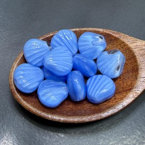 May include: A collection of light blue, shell-shaped glass beads with a ridged texture, displayed in a small wooden scoop. The beads have a translucent quality, and the wooden scoop has a natural wood grain.