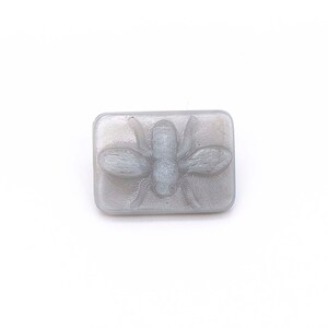 May include: A gray rectangular button with a raised, detailed image of a fly.